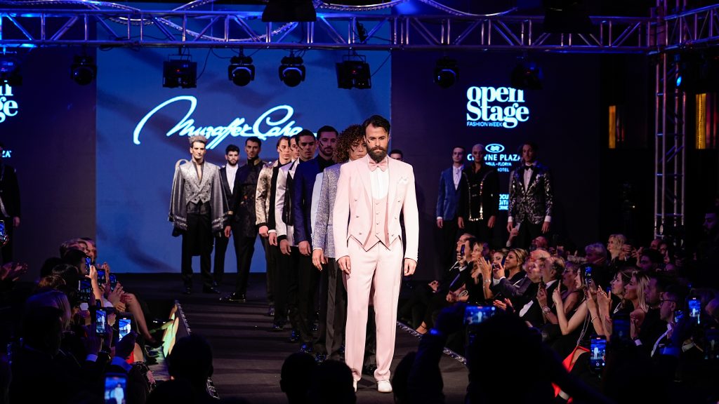Open Stage Fashion Week podyumunda sıra halinde yürüyen erkek modeller, sahne ışıkları ve izleyici siluetleri